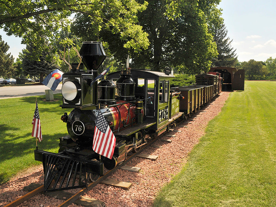 Miniature train on tracks with American flags and a Colorado flag in a park setting.